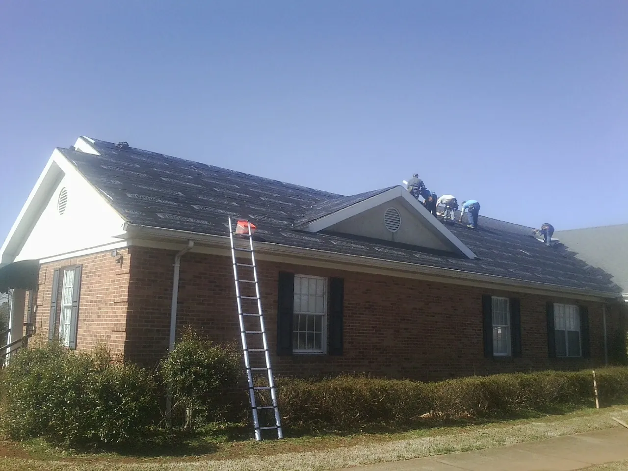 Crew tearing shingles off a brick ranch roof with a ladder set — Emergency Roof Repair work by our crew in Cornelia