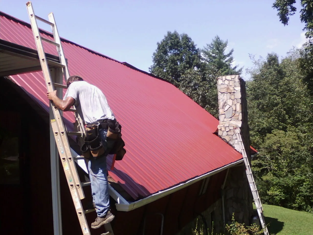 Roofer on a ladder beside a red metal roof and stone chimney — Soffit & Fascia Repair by Roofing Brothers in Cornelia